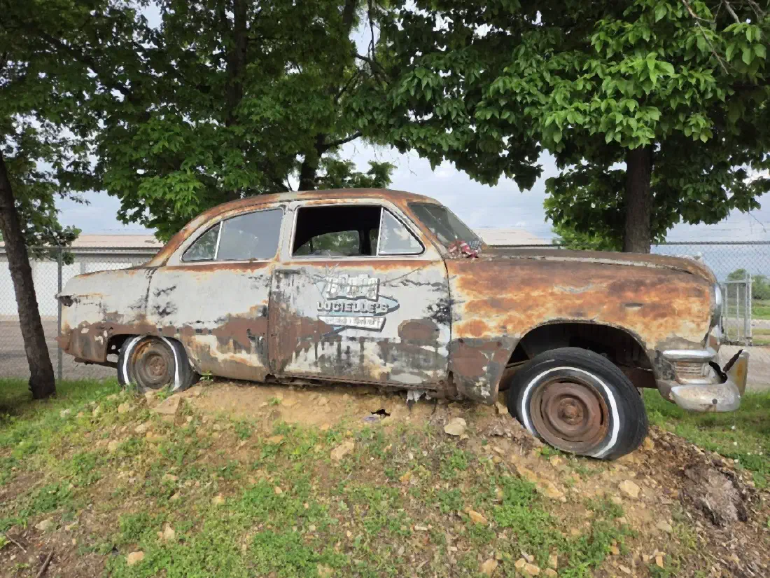 A rusted 1950s sedan with the Edith Lucielle's logo on the door, parked on a mound out front